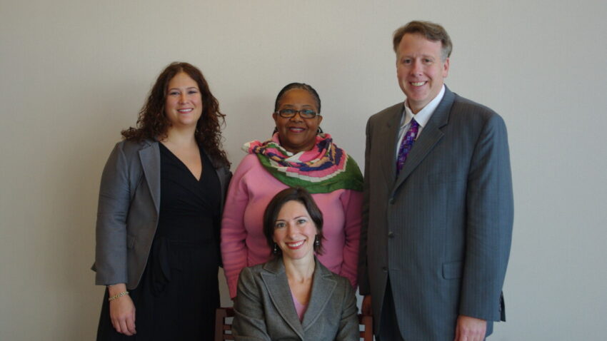 Three people -- two women and one man -- stand behind a seated smiling woman.