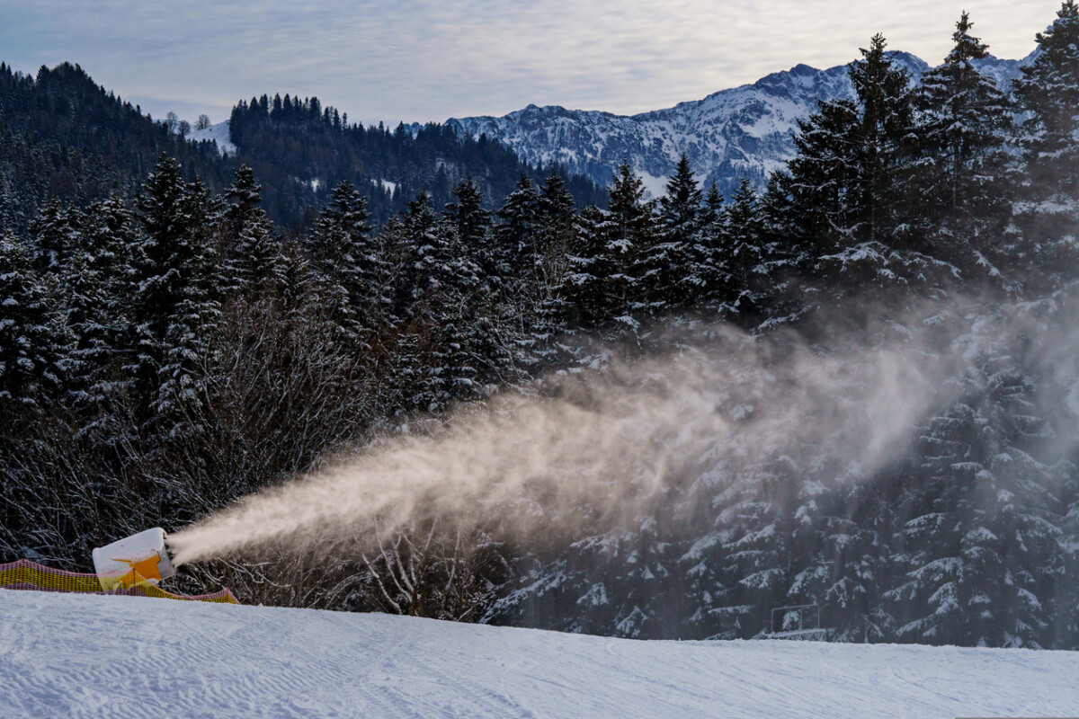 Snow cannon in operation for the production of artificial snow in the ski area. Snow fountain in front of snow-covered conifers. The Kaiser Mountains in the background