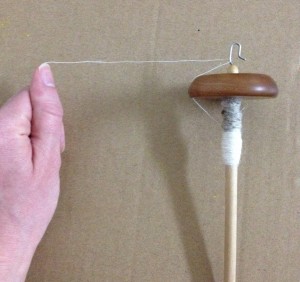 My hand holds a partially spun thread which is passed through the hook of the drop spindle on the right. Both are resting against a cardboard background.