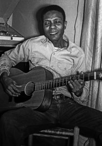 Black and white photograph of George Henry Bussey holding a guitar and singing with a smile.