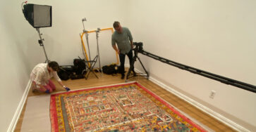 A large colorful carpet is in the foreground of the picture. Two people are standing on the side, surrounded by lighting and camera equipment