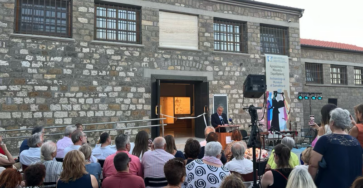 Picture taken during the Samothrace Museum opening. Taken outside where the audience is sitting on chairs listening to a speaker at the podium, this picture shows a white ribbon tied between the museum's open doors.