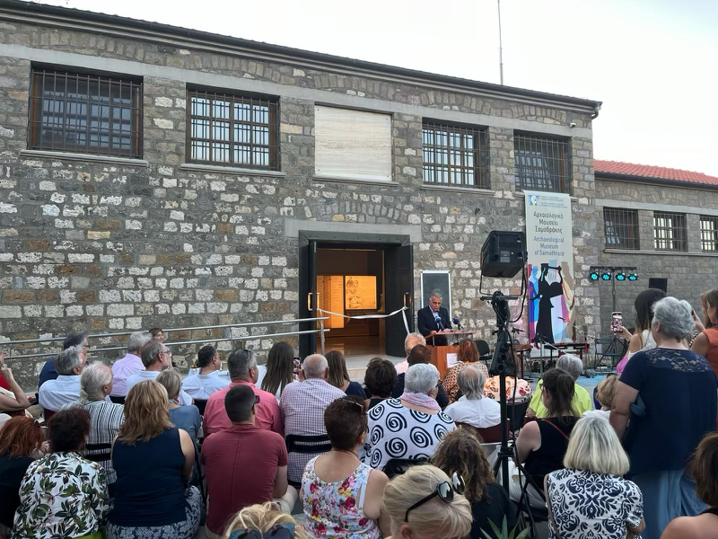 Picture taken during the Samothrace Museum opening. Taken outside where the audience is sitting on chairs listening to a speaker at the podium, this picture shows a white ribbon tied between the museum's open doors.