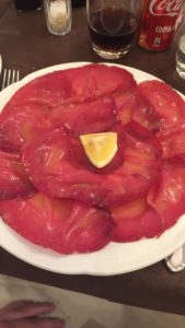 photo of thinly sliced meat on a plate. grease gathers on the meat and a lemon wedge rests on top. end alt text.