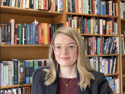 A white woman with blonde hair and clear glasses sitting in front of a bookshelf.