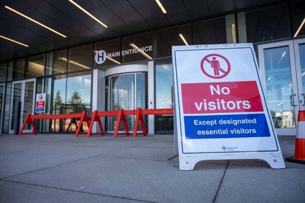 A hospital main entrance in Alberta, CA with a sign reading "No visitors; except designated essential visitors"