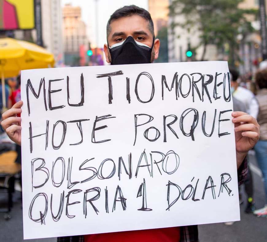 Homem em um protesto com sinal que diz "meu tio morreu hoje porque Bolsonaro queria 1 dólar"