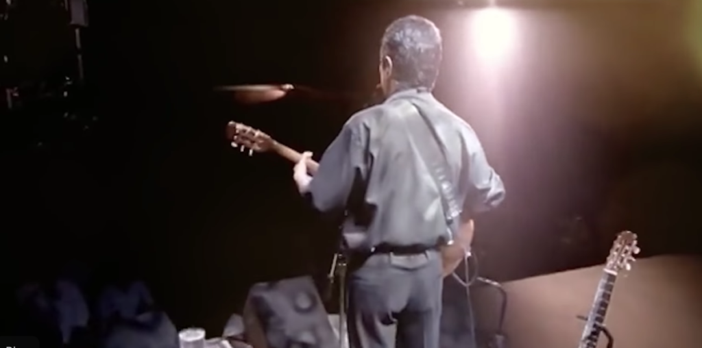 Chico Buarque de pé em um palco, com as costas voltadas para a câmera. Esta imagem foi usada no vídeo do Leite.