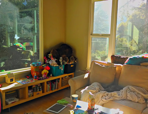 A messy living room with beautiful afternoon light streaming in through windows and children's toys piled on a shelf in the corner