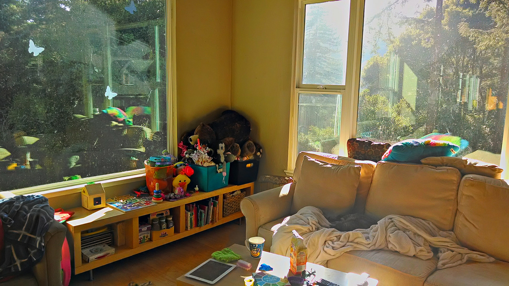 A messy living room with beautiful afternoon light streaming in through windows and children's toys piled on a shelf in the corner