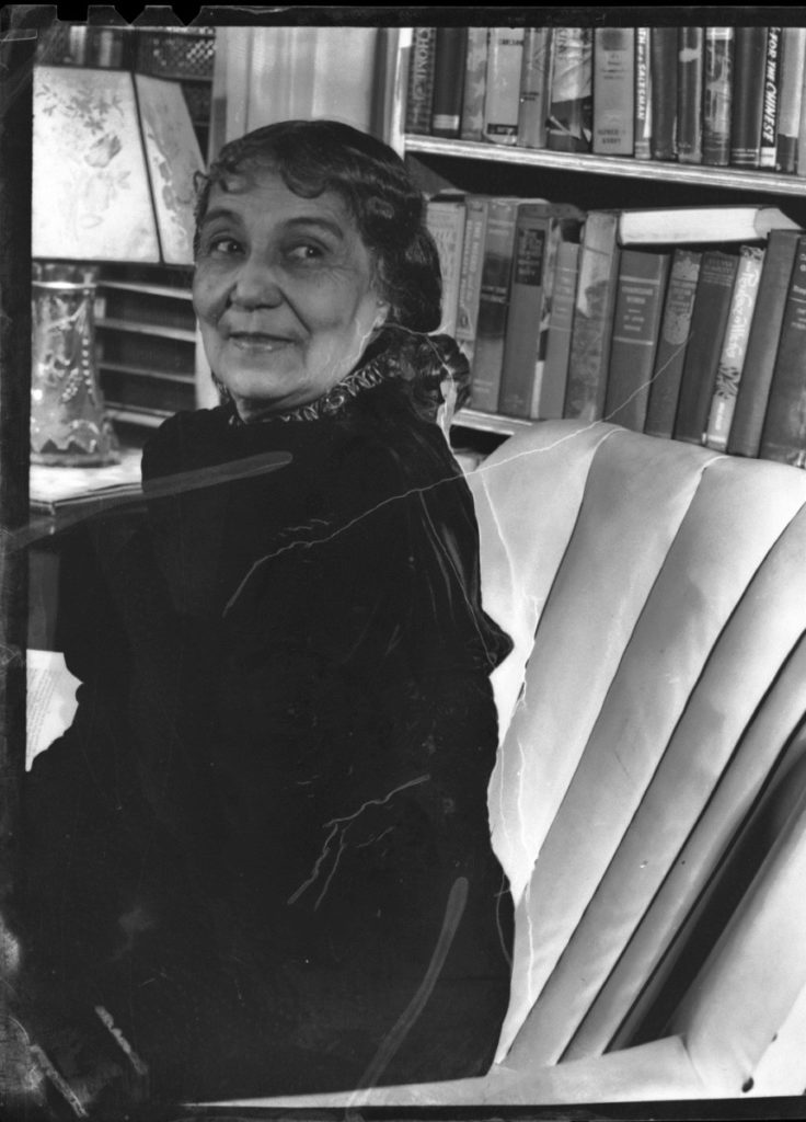 Photo of Anne Spencer in old age, sitting in a chair in front of her bookcase.