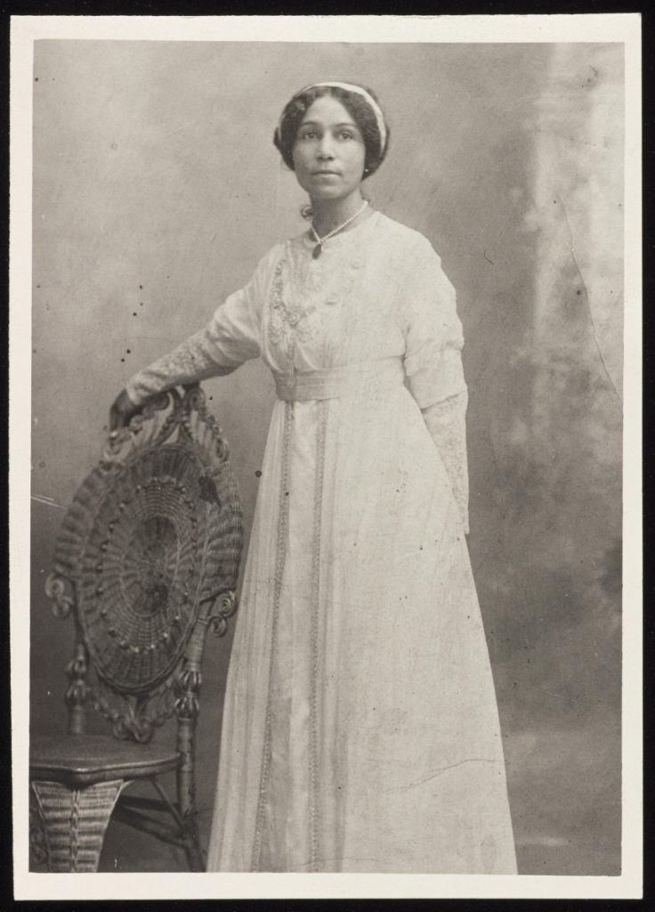 Young woman in a wedding dress standing for a photo with her arm draped over a chair