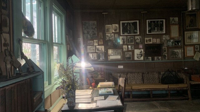 A desk with lamp facing out a window. Lots of photographs on the wall to the right of the desk.