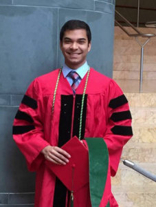 Dhruv Amratia smiling with short dark brown hair and wearing a red scholar's robe with black velvet detailing, holding a red four-cornered graduation cap and red and green hood. Underneath is a light blue colored shirt and patterned tie.