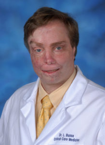 Headshot of Laurence Busse smiling with short, dark blonde hair, wearing a white labcoat emblazoned with Dr. L. Busse, Critical Care Medicine in blue above the left breastpocket. Underneath is a light yellow collared shirt and slightly darker yellow tie dotted with light blue.