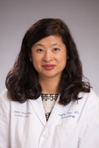 Headshot of Suephy Chen smiling with medium length curly black hair, wearing a white lab coat with text Emory Healthcare on right side and Suephy Chen, MD, Dermatology in blue script above left breastpocket. Underneath is a patterned white shirt with black embroidery.