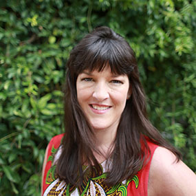 Headshot of Dabney Evans against a backdrop of green leaves, smiling with medium length brown hair and bangs/fringe, wearing a red shirt patterned with green and white embroidery.