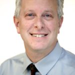 Headshot of Scott Fridkin smiling with medium short gray hair, medium gray shirt, and dark patterned tie.