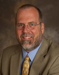 Headshot of Robert Hampton smiling, with glasses and a beard, wearing a khaki colored blazer, light blue collared shirt, and patterned gold tie.