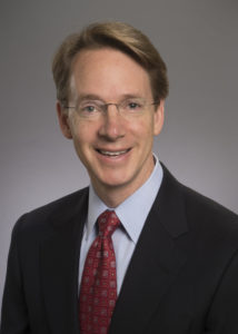 Headshot of Gregory Martin smiling with short blonde-ish hair and glasses, wearing a dark colored blazer, light blue colored shirt, and dark red patterned tie.