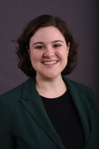 Headshot of Emma Rary smiling with short curly brown hair, wearing a dark green blazer and black shirt.