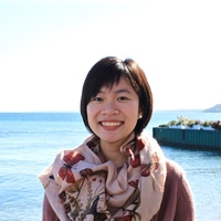 Headshot of Milkie Vu smiling against waterscape backdrop, with short black hair, wearing a patterned beige scarf, pearl earrings, and light colored sweater.