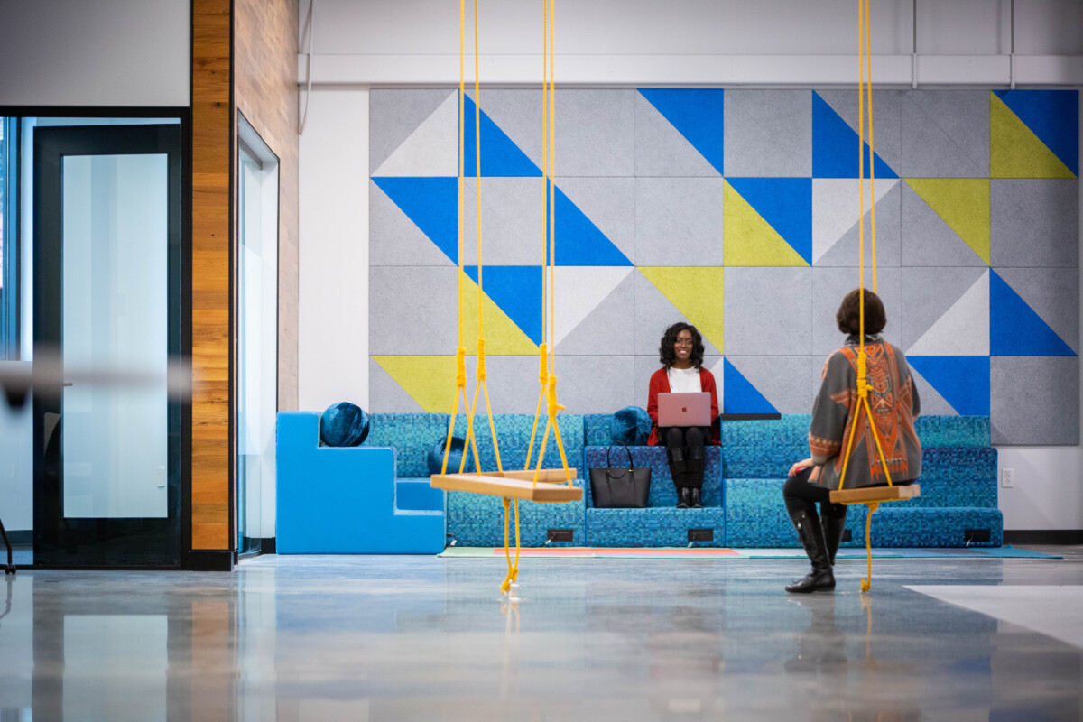 Two women sit in an open, colorful industrial space. One woman is on a blue couch with a laptop, and the other sits on a swing with her back facing the camera.
