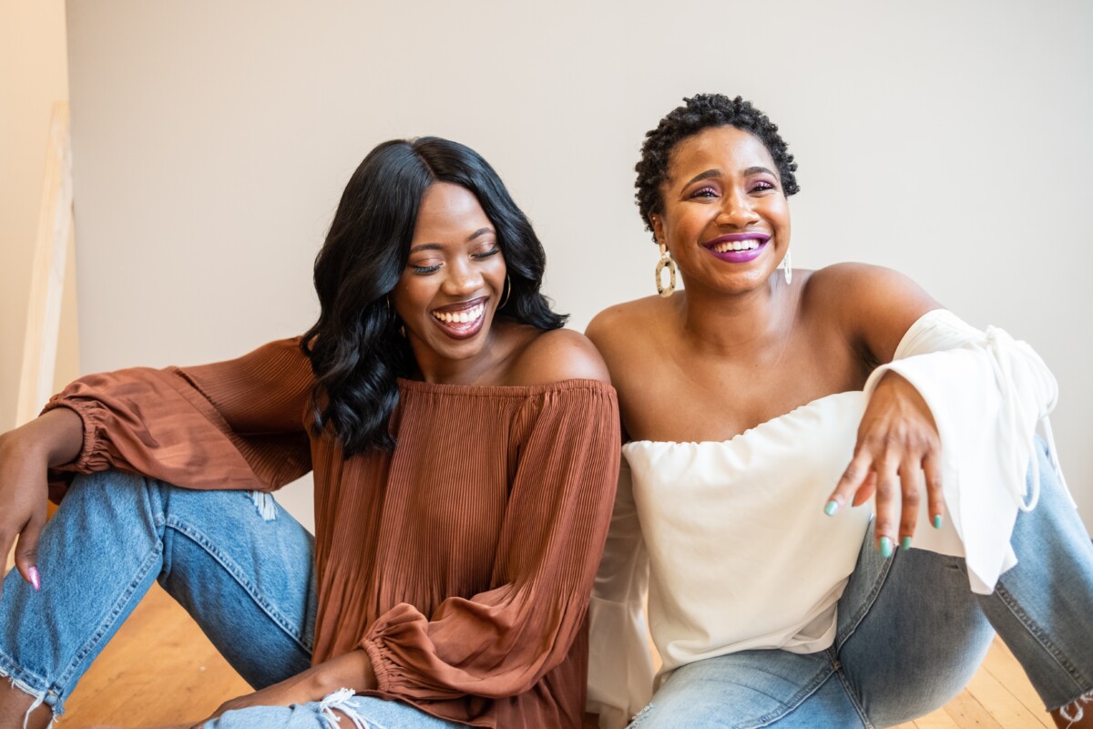 Two Black women sit on the floor and lean on each other, smiling