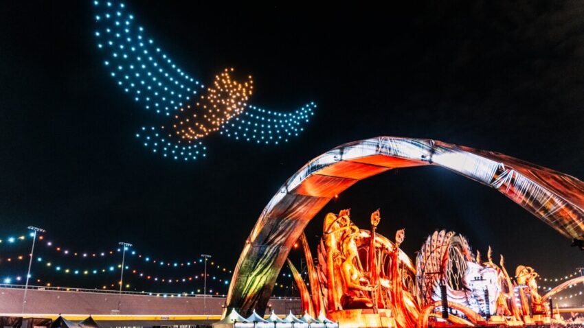 Lighted blue and orange drones create the shape of an owl in flight above a music festival.