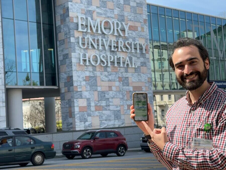 Santiago Arconada Alvarex stands outside Emory University Hospital, holding his phone and smiling to the camera