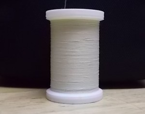 a sool of white commercial thread resting on a wooden surface against a black background. The spool has a needle sticking out of it.