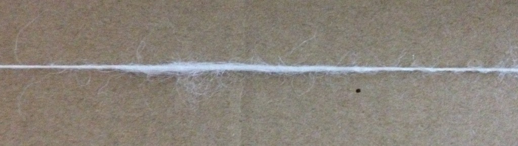 Apiece of my handspun thread with a section in the middle much thicker than the rest. The thread is resting against a cardboard background
