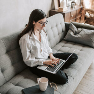 Girl in white blouse sitting on gray couch working on laptop computer. Image credit: Vlada Karpovich/Pexels