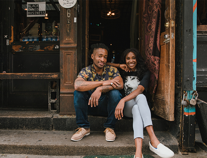 Two black cafe owners, one male and onw female, sit on the steps outside their business on a downtown street. A sign hanging from the cafe window says "black owned business."outside