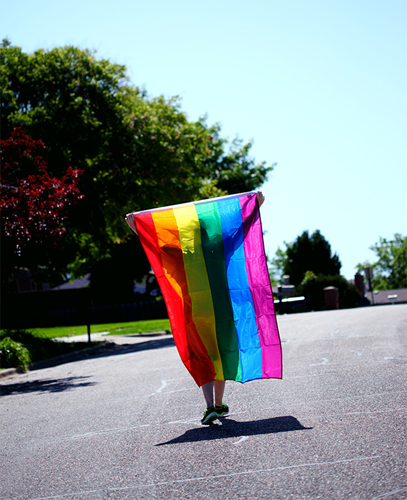 Person shown in silhouette behind a rainbow flag, walking across a parking lot in bright sunlight.