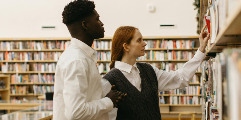 Two people look at a shelf of books in a library.