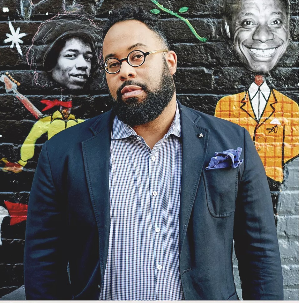 African American poet Kevin Young, dressed in a collared shirt and open blazer, stands in front of a mural depicting African American artists.